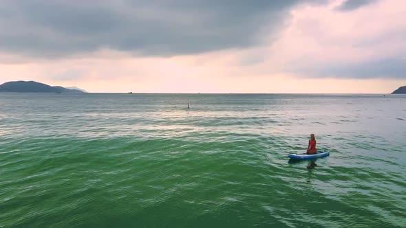 Backside Girl Sits on Paddleboard in Ocean Against Seashore