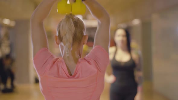 Back View of Blond Caucasian Sportswoman Lifting Weights with Blurred Brunette Personal Trainer