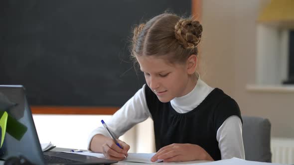 Smart Confident Teenage Schoolgirl Watching Online Lesson Writing with Pen Sitting at Desk Indoors