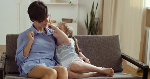 Two People Caucasian Mom Caressing Touching Noses with Cute Kid Daughter Sitting on Sofa at Home