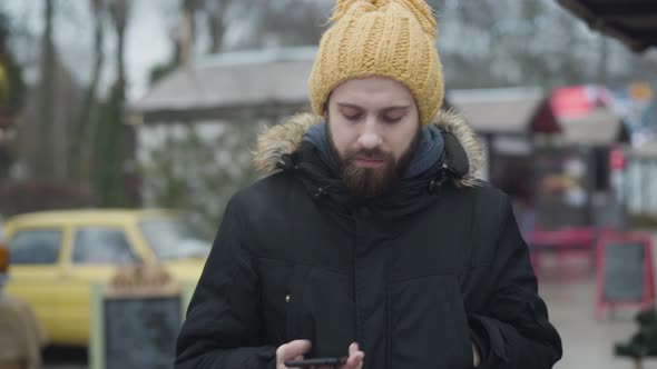 Young Frozen Caucasian Man Standing Outdoors Dressed in Warm Clothes and Checking Time on Smartphone