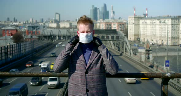 Young Man Is Putting Surgical Mask on Face Standing Outdoors at City Bridge Over Road, Epidemic