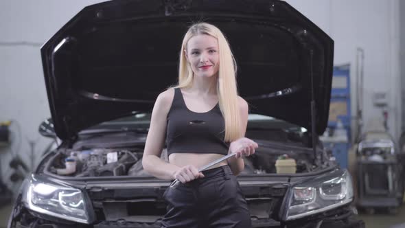 Sexy Blond Caucasian Woman Standing at the Background of Open Car Hood with Adjustable Wrench