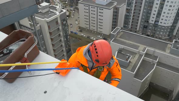Industrial Climber in Orange Suit and Helmet Prepares To Descend From Roof of Multi-storey Building