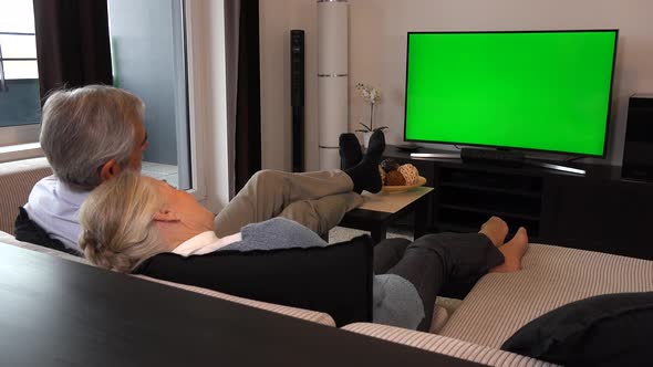 An Elderly Couple Sits in A Living Room in An Apartment and Watches TV with A Green Screen