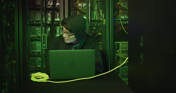 Asian male hacker in hoodie using laptop by computer servers
