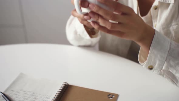 Young Pensive Girl In A White Shirt Drinking Morning Coffee While Sitting At A White Table, Planning