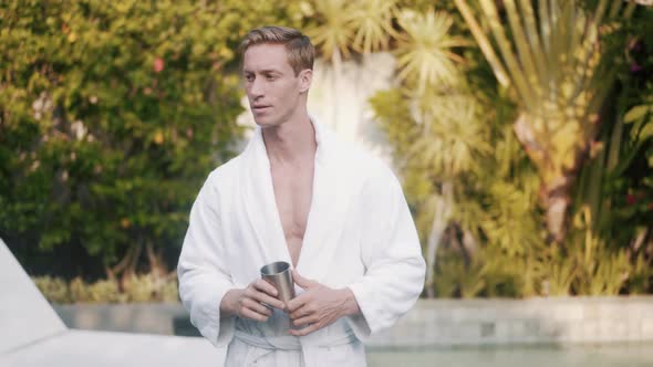 Man in Bathrobe Drinks Coffee in Early Morning Outdoors, Greenery on Background