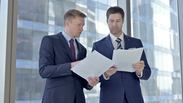 Two Middle Aged Businessmen Checking Documents Against Boardroom Office Window