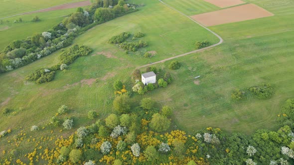 Brick hut sitting at the end of a countryside dirt road surrounded by lush fields and blossoming bus