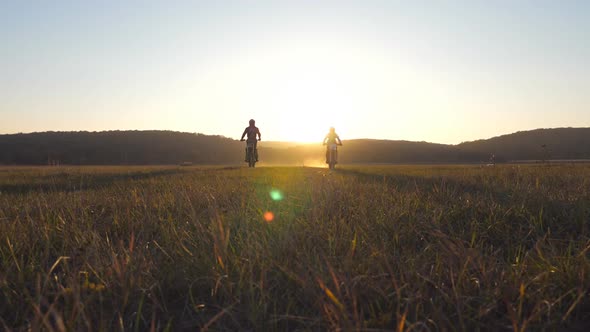 Two Motorcyclists Passing Through Large Field with Beautiful Sunset at Background