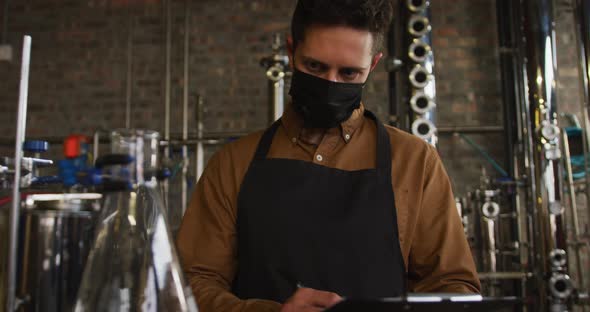 Portrait of caucasian man working at gin distillery wearing face mask and apron looking to camera
