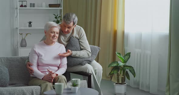 Elderly Caucasian Couple Sits on Gray Sofa Old Man Loving Husband Hugs His Wife Comforts Beloved Sad