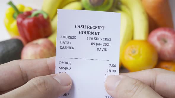 Hands hold shopping receipt bill with variety of colorful fresh fruits and vegetables.