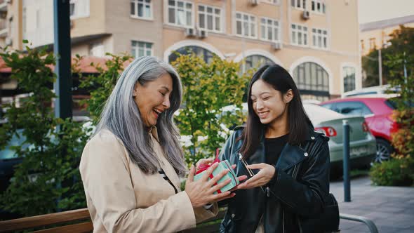 Asian Daughter is Taking Out Gift Box From Paper Package and Giving It to Her Surprised Mature Mum