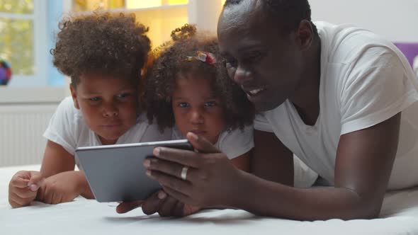 African American Father and Preschool Children Lying Together on Bed and Using Digital Tablet