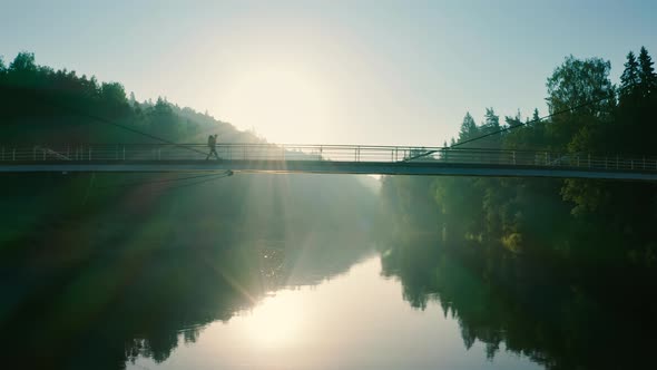 Profile shot: single adult individual crossing a bridge over a wide sunlit river, aerial slider