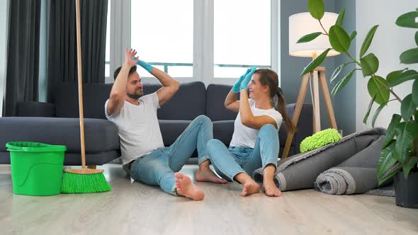 Tired Couple Sits on the Floor in a Room and High Five Each Other After They Finish Cleaning the