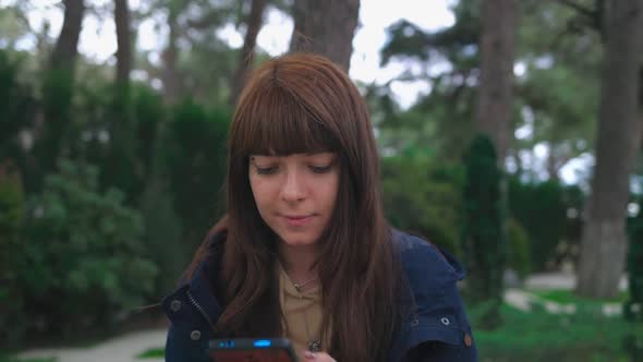 A Young Woman Sits on a Park Bench and Types on Her Smartphone