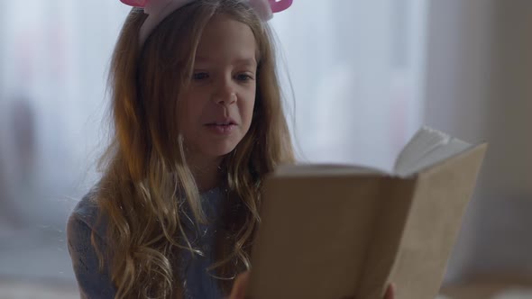 Portrait of Charming Happy Little Girl Reading Out Loud Sitting at Home Indoors