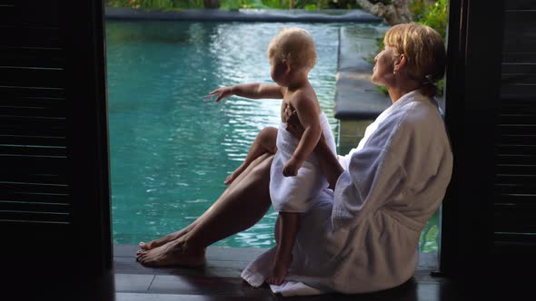 Grandmother Sitting on the Edge of the Swimming Pool with Her Grandchild in a White Robe