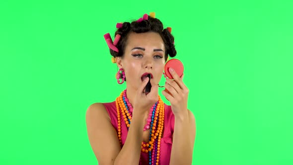 Girl with Curlers on Her Head in a Pink Dress Looking in Red Mirror and Paints Her Lips. Green