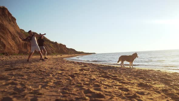 Young Beautiful Couple Throwing Stones in Water Walking with Dog at Seaside at Sunrise