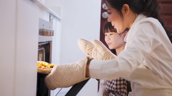 Asian young mother and daughter open the oven and bring pizza from machine in kitchen at home.