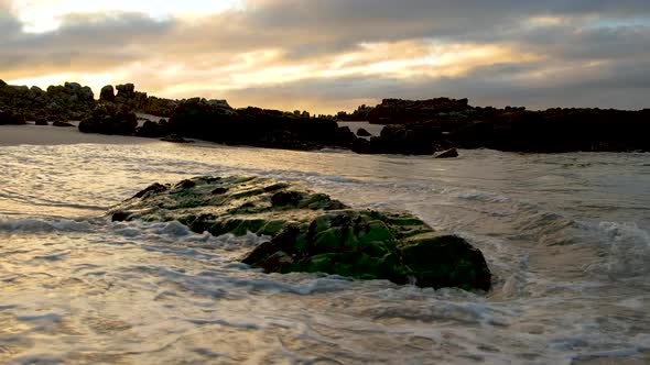 Low waves breaks around wet rock on beach, low angle shot during sunrise, golden reflections and sof