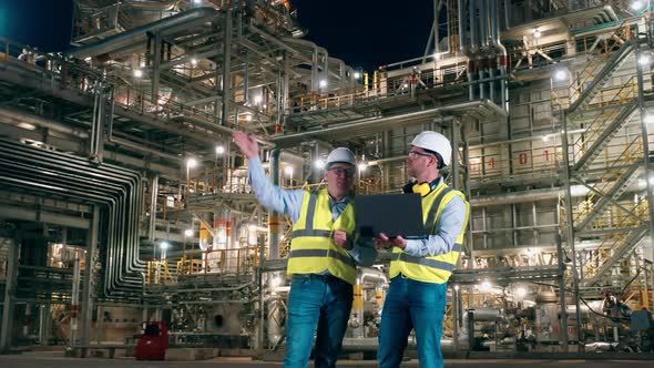 Two Engineers with a Laptop are Talking Inside of the Oil Refinery