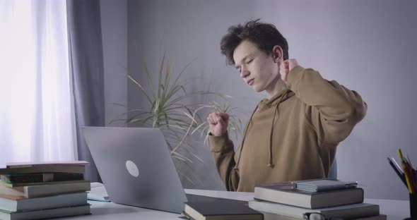 Teenage Boy Stretching As Sitting at the Table with Laptop and Books. Portrait of Tired Caucasian alt