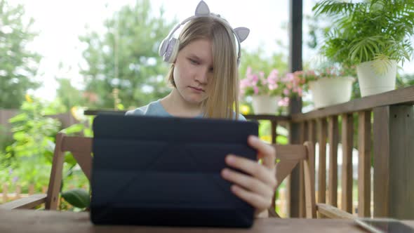 Teenage Girl Sits At Wooden Table In a Summer Cafe In Headphones With Phone