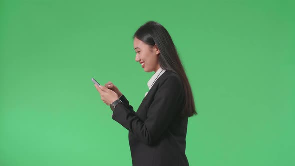 Side View Of A Smiling Asian Business Woman Using Mobile Phone While Walking On Green Screen