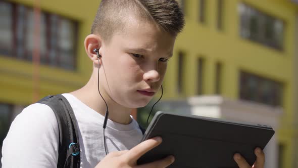 A Caucasian Teenage Boy Works on a Tablet with Earphones on  Closeup  a School in the Background