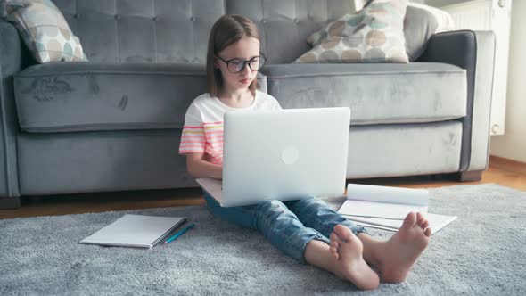 Serious 8 Years Old Girl in Glasses Typing on a Laptop While Sits on a Carpet