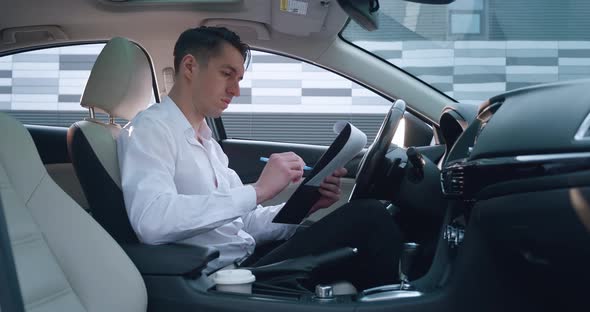 Portrait of Serious Male Entrepreneur Sits on Driver's Seat in Car Reads and Signs Paper Documents