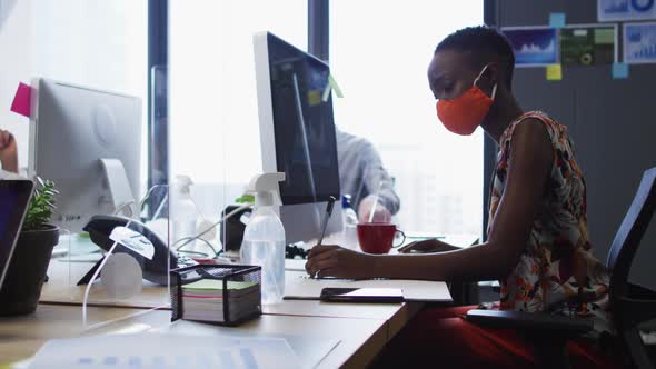 African american woman wearing face mask taking notes while sitting on her desk at modern office
