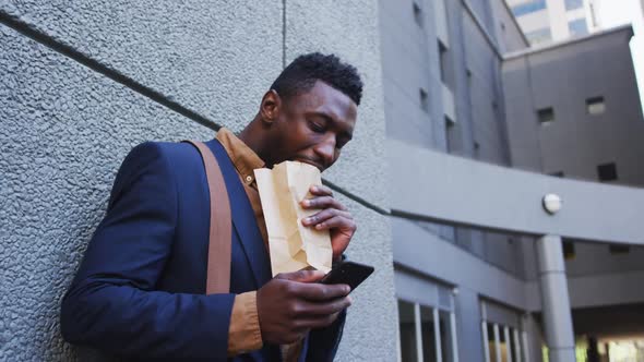 African american businessman eating sandwich using smartphone in city street