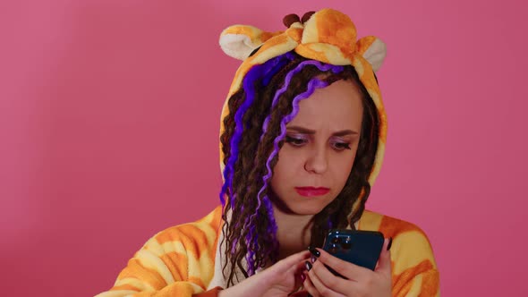 Young Cute Disappointed Woman with Dreadlocks in Kigurumi Browsing Mobile Phone on Pink Background