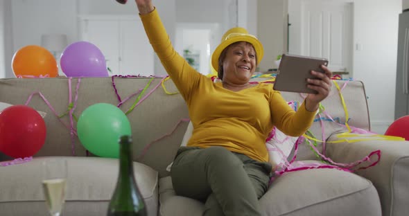 Happy african american senior woman using party blower making new year's eve tablet video call
