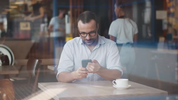 View Through Window of Young Man Sitting in Cafe Using Mobile Phone