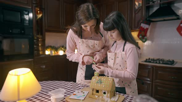 Charming Caucasian Girl Placing Santa on Decorated Gingerbread House Helping Woman Cooking Dessert