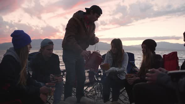 Man lifts kettle out of campfire to fill people mugs at sunset