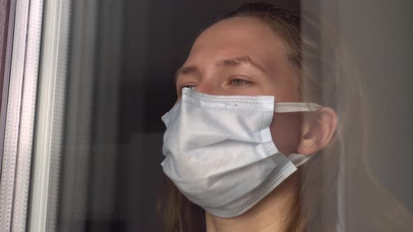 A young woman in a medical protective mask looks out the window