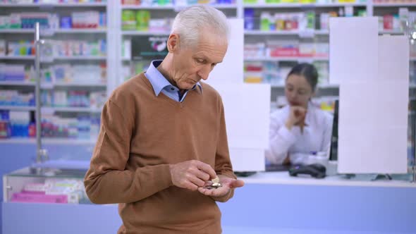 Sad Poor Senior Caucasian Man Counting Coins Standing in Pharmacy Looking Back at Cash Desk with
