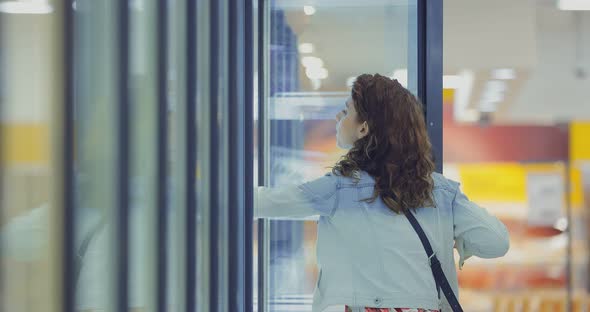 Young Beautiful Woman Takes Frozen Vegetables From a Refrigerated Supermarket Display Case