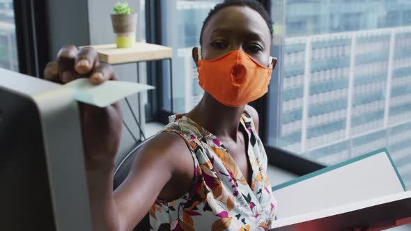 African american woman wearing face mask reading memo note while sitting on her desk at modern offic