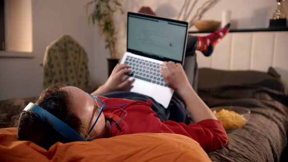 Young Man Lying on the Bed and Searching the Internet and Eating Chips