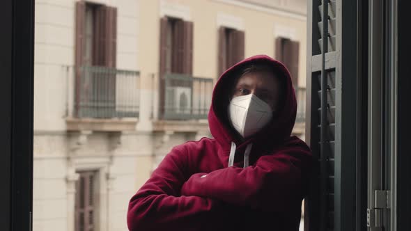 Portrait of Millenial Man in Hoodie and Protection Mask on Balcony Near Window Spain Barcelona