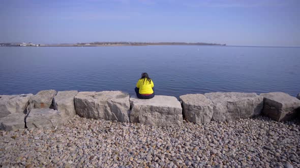 Person in Toronto Trillium Park with headphones sitting near lake in bright yellow jacket during day